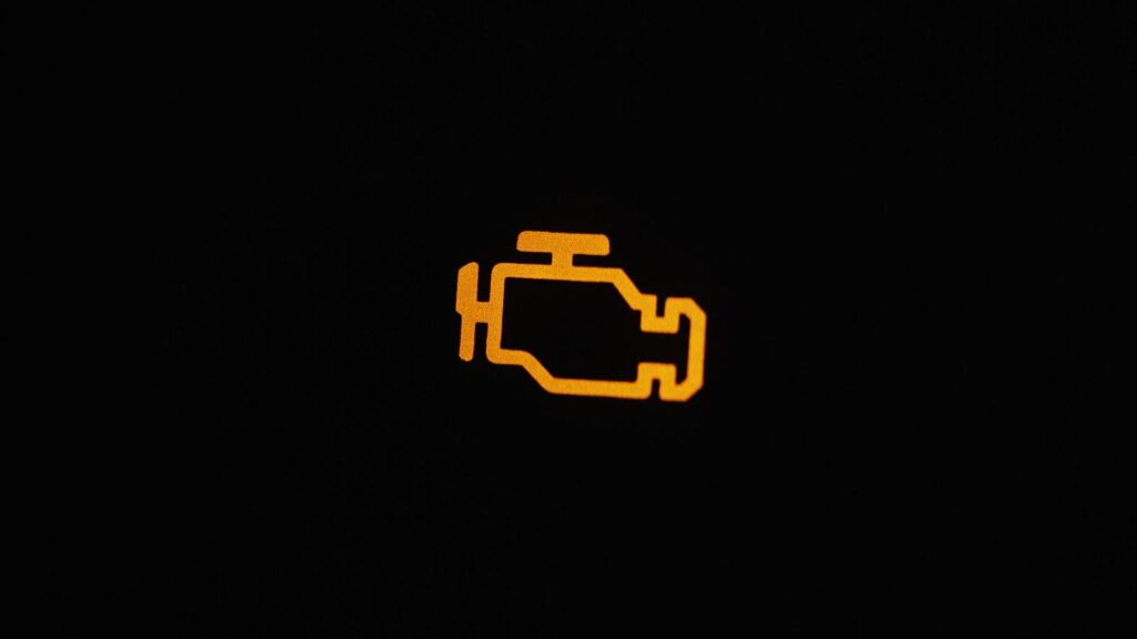 An illuminated check engine light on a dark dashboard. The light shows an engine block icon, signaling a potential issue that should be inspected by a mechanic at America 1st Automotive in Stafford, Virginia.