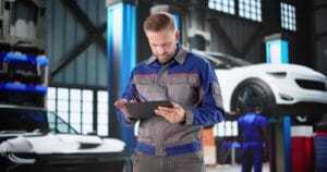 Male mechanic in a blue-gray uniform uses a tablet in a car repair shop with cars on lifts behind him.