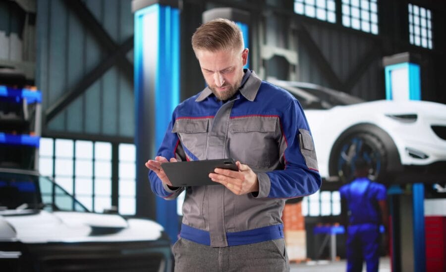 Male mechanic in a blue-gray uniform uses a tablet in a car repair shop with cars on lifts behind him.