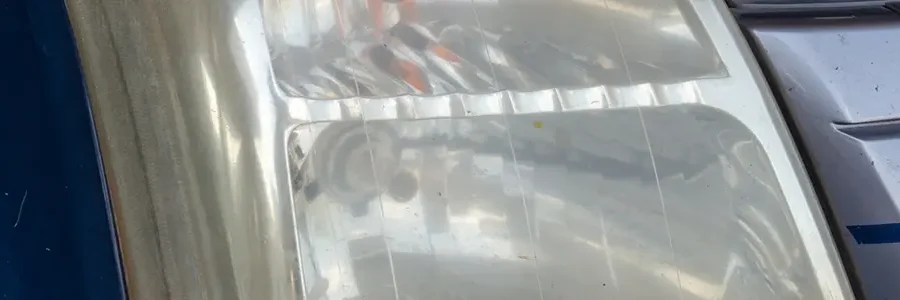 Close-up of a car's headlight and surrounding bodywork, showing a clear plastic lens and reflections on a metallic surface.