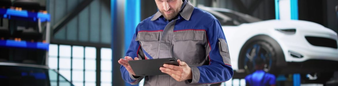 Male mechanic in a blue-gray uniform uses a tablet in a car repair shop with cars on lifts behind him.